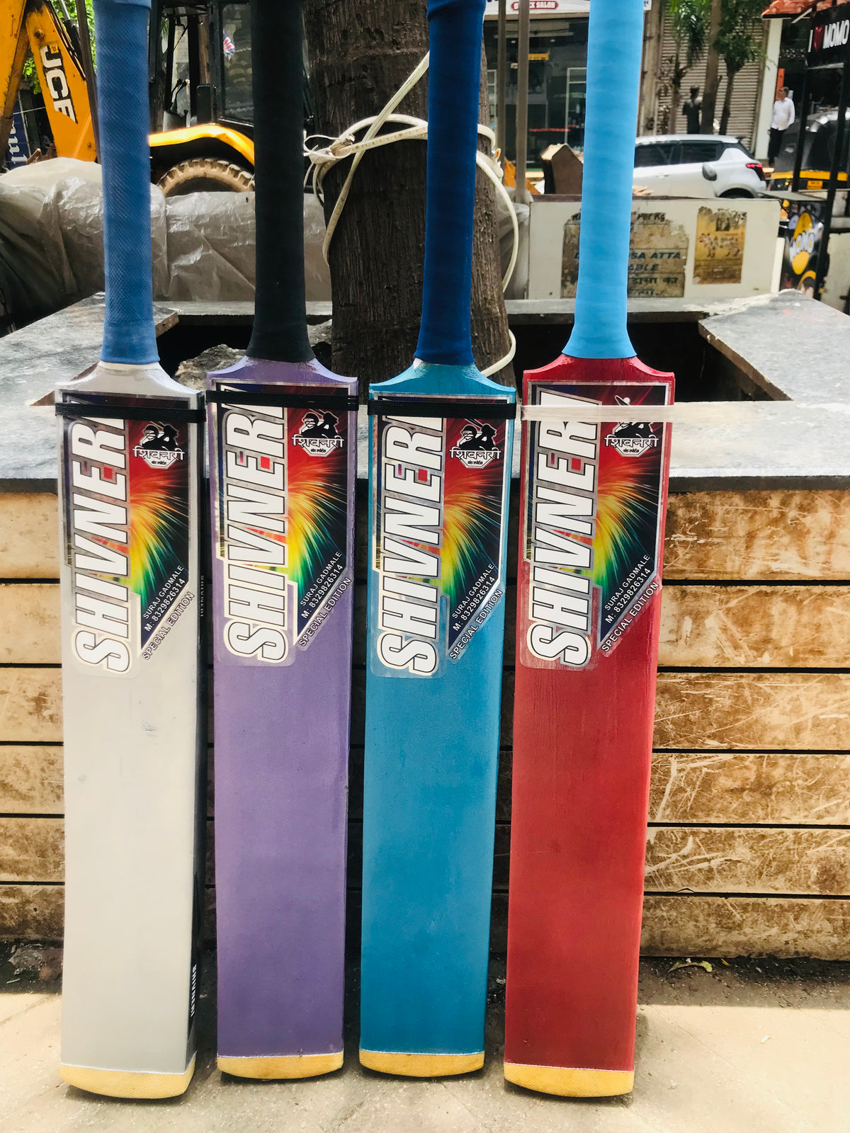 Four colorful cricket bats with 'Shiv Naresh' branding on a wooden surface.